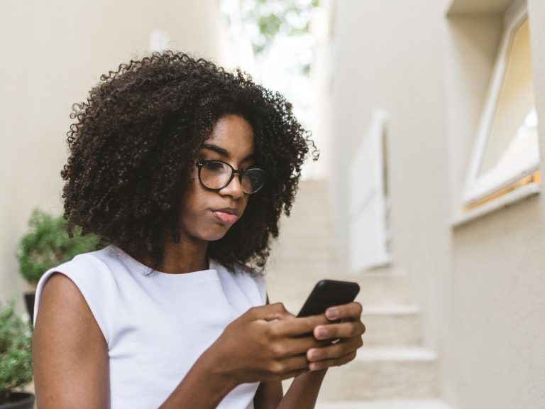 Young woman with curly hair and glasses using a smartphone, sitting on stairs, reflecting on pregnancy-related services and resources.